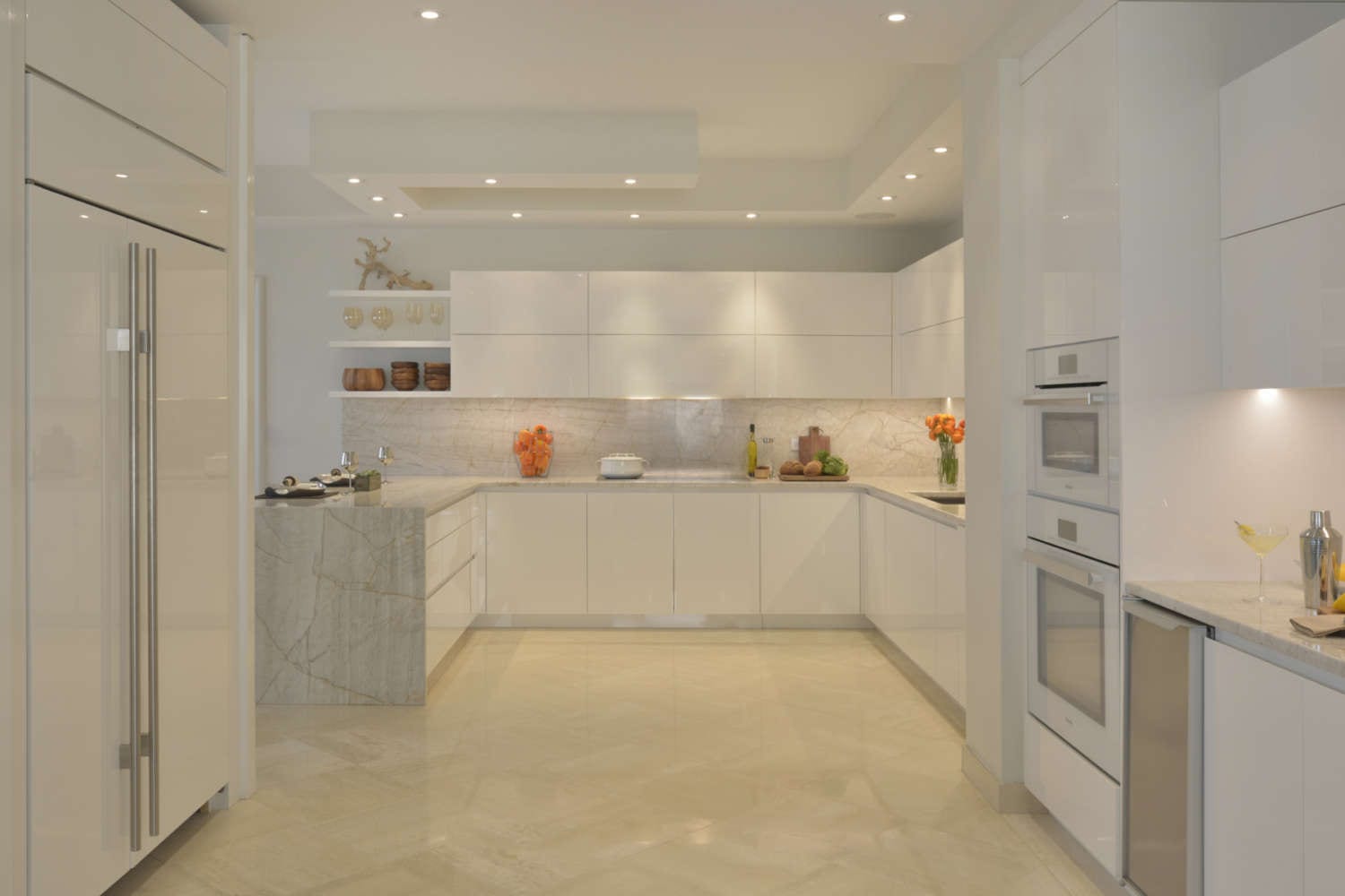 U-shaped kitchen is designed with soft lighting and a palette of warm whites. The fully custom cabinets are ArtCraft flat panel, frameless in high gloss white, countertops and backsplash are light marble with veining, and the floor is a light porcelain tile. Open shelving is used for display.