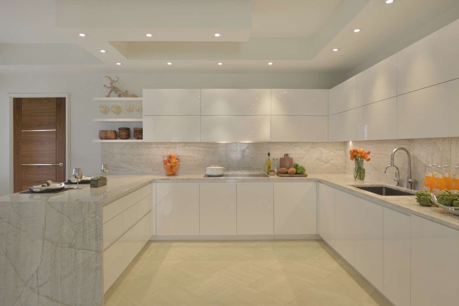 U-shaped kitchen features high gloss white, flat panel fully custom Artcraft cabinetry, light marble countertop and backsplash with veining, light porcelain tile flooring, open shelving and soft lighting. Design by Tabitha Tepe of Bilotta Kitchens.