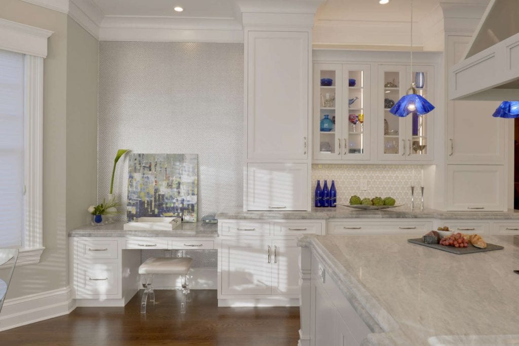 Bright U-shaped kitchen with white painted fully custom Bilotta cabinets, light marble countertops, and desk in kitchen. Cabinet fronts are a mix of full panel and glass front doors. Designed by Fabrice Garson of Bilotta Kitchens.