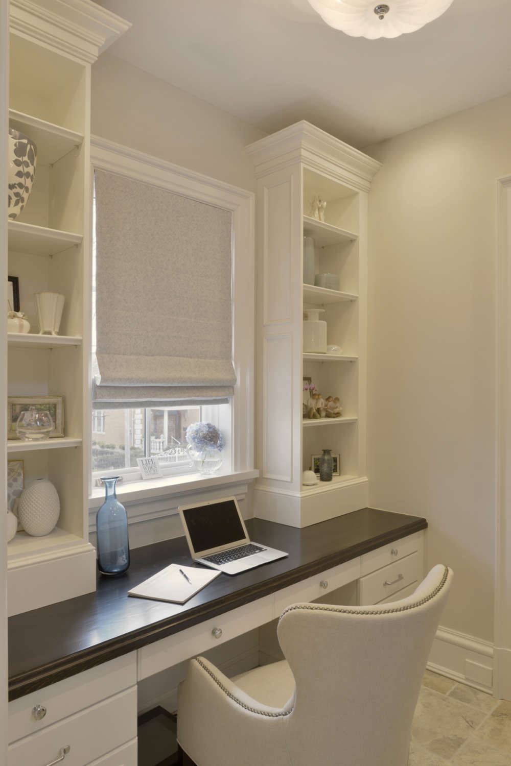 Home office features a classic white painted, custom desk with round polished stainless hardware and espresso brown top. The desk is positioned under a window with flax colored roman fabric shade flanked by custom white painted built-in open shelving.