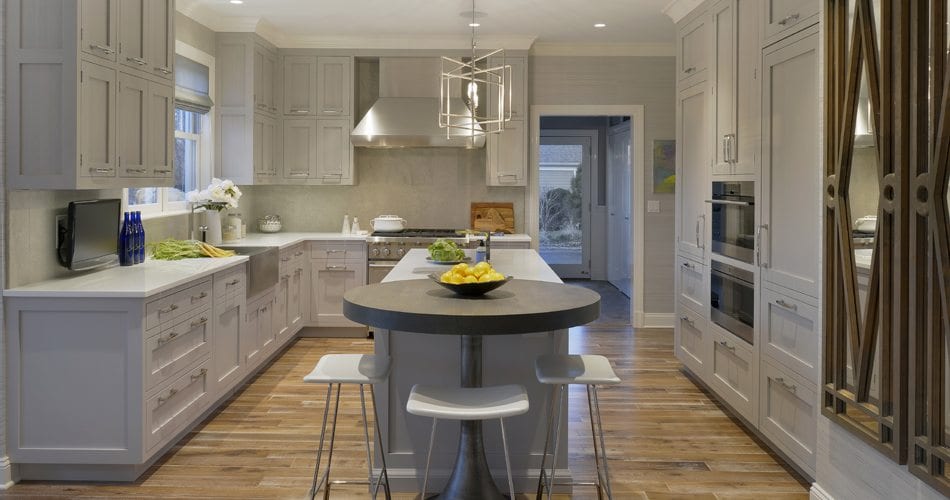 U-shaped kitchen with breakfast nook features coffered ceiling with silver-accented pendant lighting, Gray granite countertop, and white painted fully custom Bilotta cabinetry with silver hardware. Upper cabinets are lit, reflecting light to the white marble backsplash.