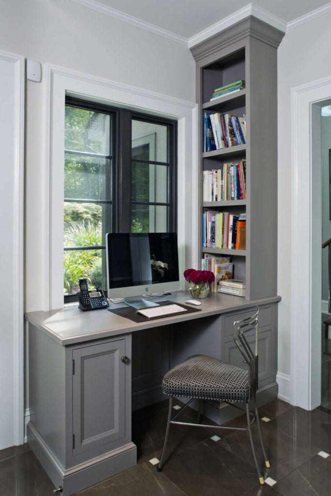 Home office features custom light grey painted Bilotta Cabinetry desk and custom built-in shelving. Desk is situated under a white-framed window with black mullions. Design by Paula Greer, CKD, of Bilotta Kitchens.