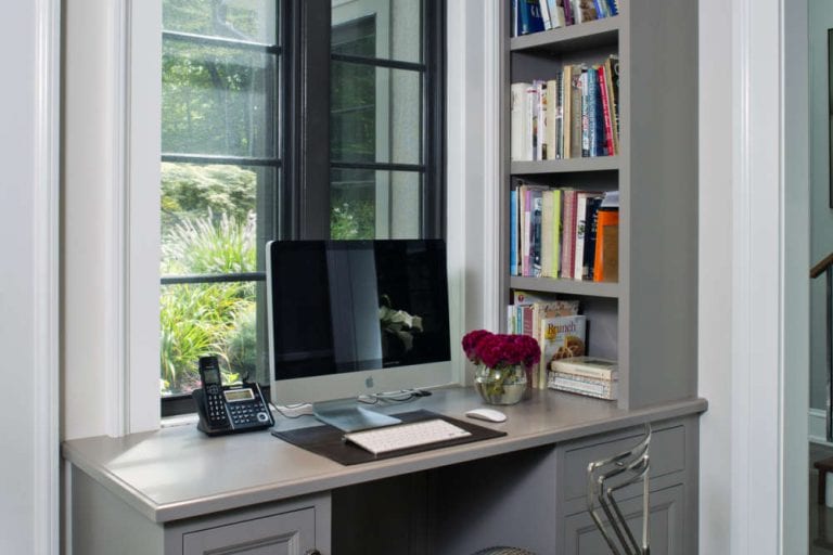 Kitchen office desk features custom light gray painted Bilotta Cabinetry desk and custom built-in shelving. Desk is situated under a white-framed window with black mullions.