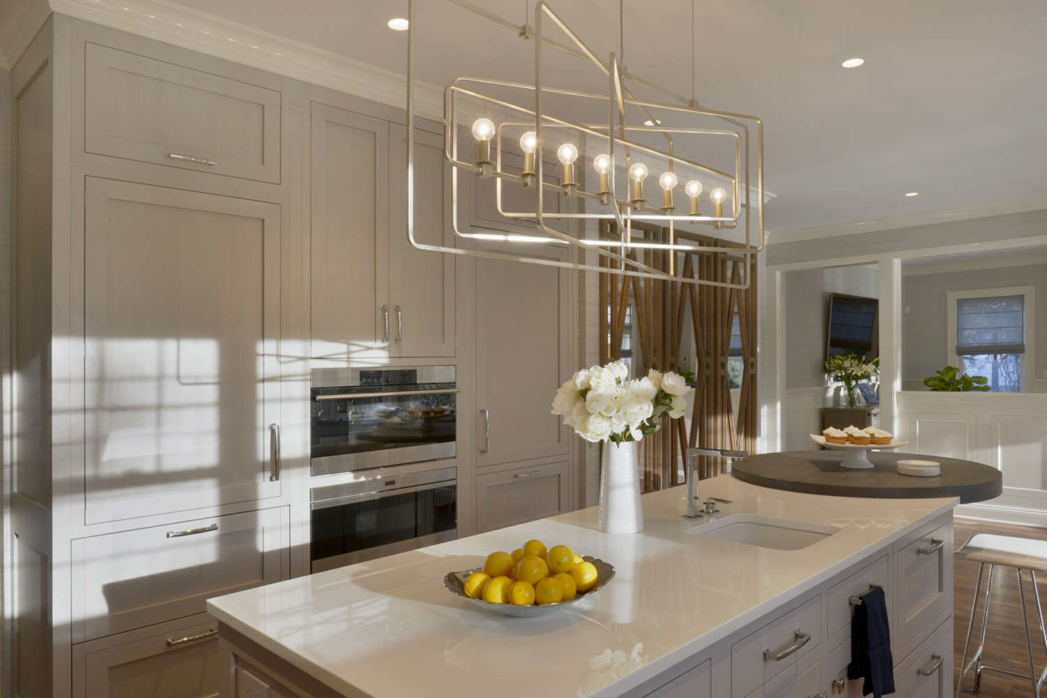 Island of open U-shaped kitchen in Rye NY features white quartz countertops, rift cut whote oak shaker style Bioltta Cabinetry, an adjacent table with seating and a dramatic gold geometric chandelier. The kitchen design by Randy O'Kane, CKD, of Bilotta Kitchens also includes fully custom shaker style white painted Bilotta Cabintry.