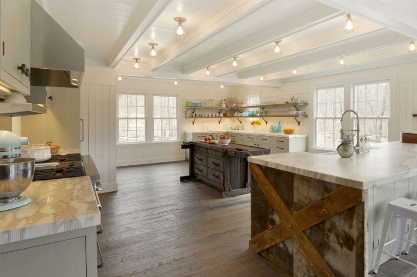 Expansive country kitchen features white beadboard ceiling and walls, exposed lighting, oversized windows, wood plank flooring, fully custom flat panel inset pale green painted Bilotta Cabinetry with oil rubbed bronze hardware, open shelving, white marble countertops and a rustic custom wood island. Design by Paulette Gambacorta of Bilotta Kitchens.