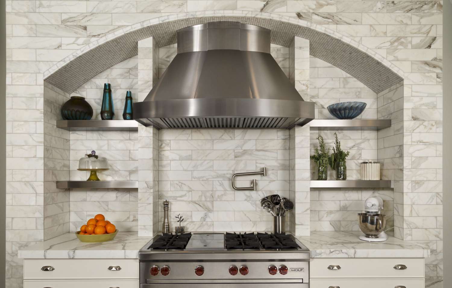 Marble brick hearth designed around stainless hood features arch and built-in stainless shelves. Range is flanked by white-painted custom kitchen cabinets.