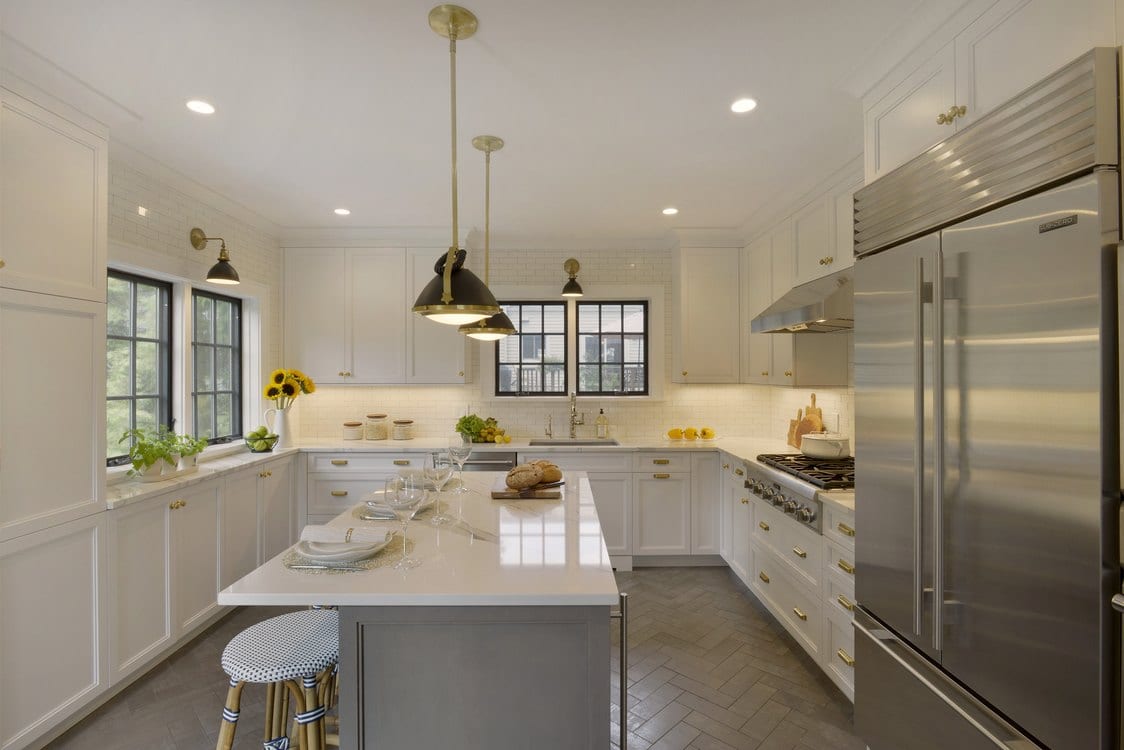 Light-filled transitional kitchen features white painted Bilotta custom cabinetry with brass hardware, glazed ceramic floor tiles set in a herringbone pattern and black and brass pendant lighting.