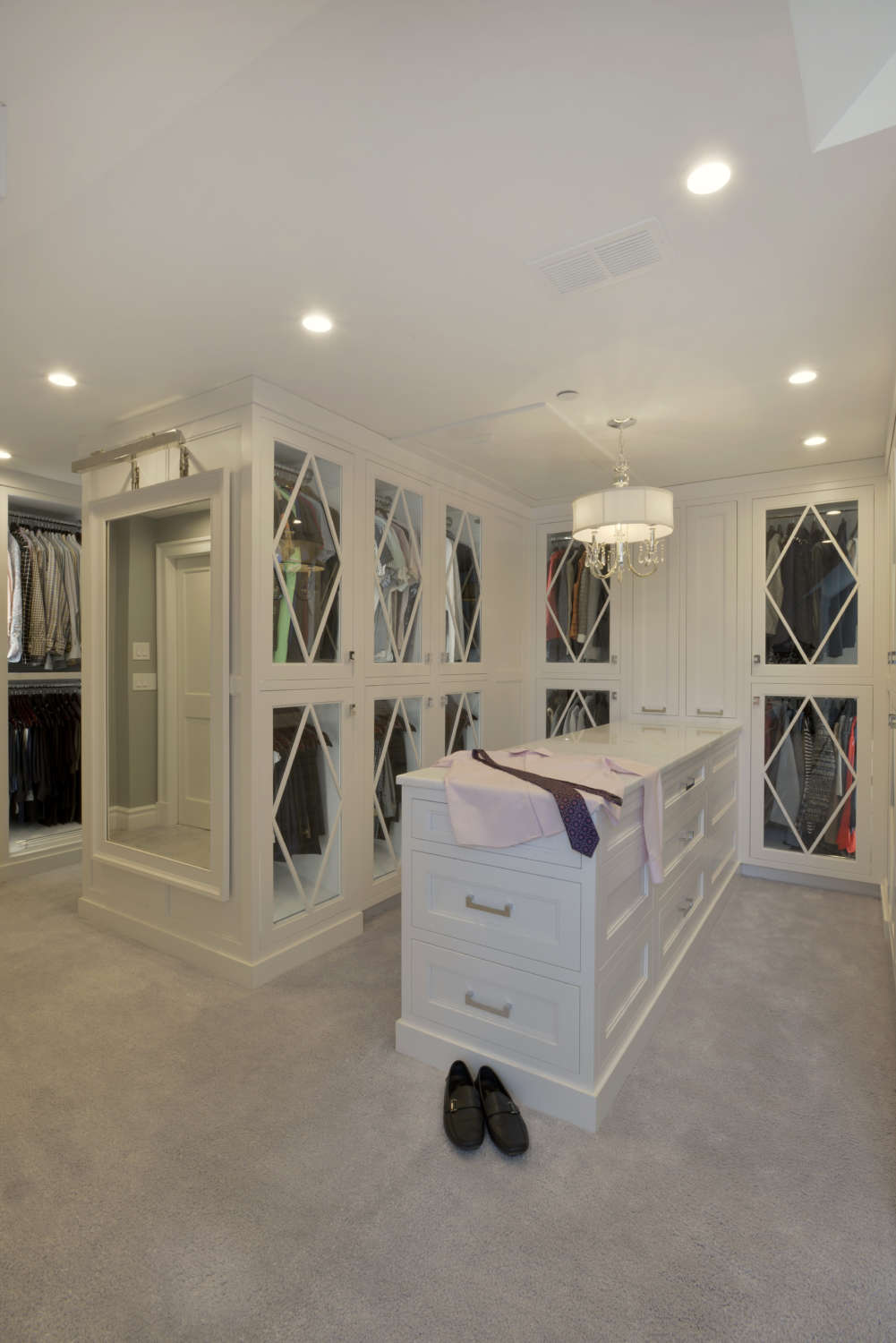 Expansive, walk-in closet outfitted with white painted custom Bilotta cabinetry with glass doors featuring decorative mullions and a marble-topped island with drawers for additional storage.