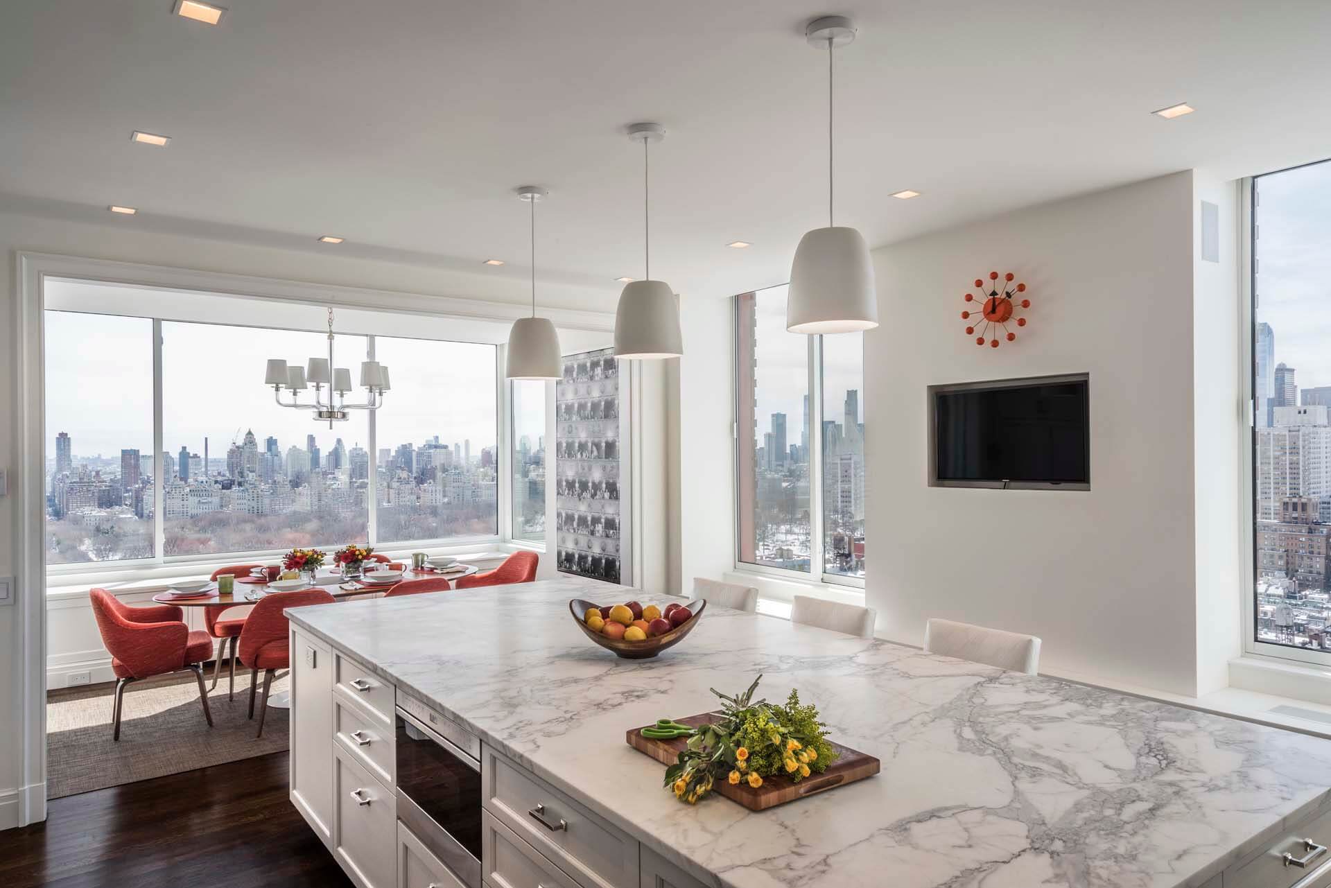 All-white kitchen features marble-topped Rutt cabinetry island with built-in microwave and an adjacent dining area with skyline view.