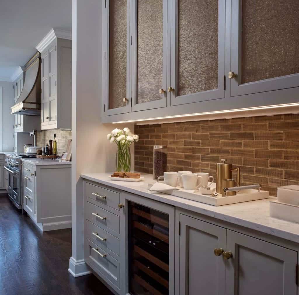 Eclectic kitchen features pale taupe cabinetry with inset doors with gold fabric inserts and variegated stone backsplash.