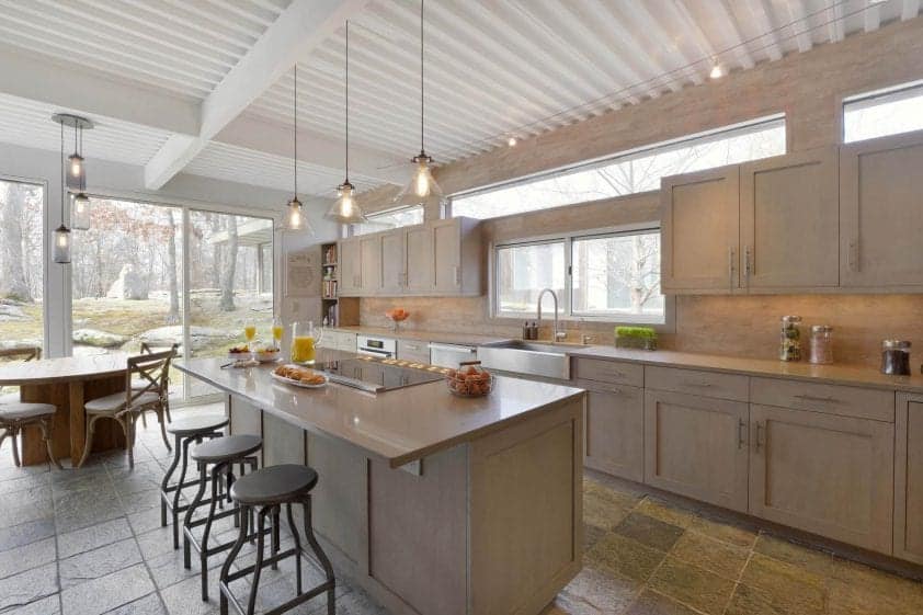 Rustic kitchen is flooded with light from sliders, transom windows and the industrial pendant lights over the island. Kitchen blends neutral quartz counters with soft travertine backspash and floor tiles and frameless, shaker style rift cut white oak Bilotta cabinets with Driftwood stain.
