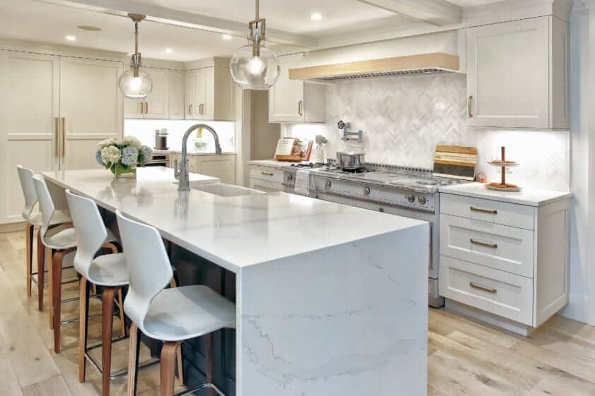 Kitchen features gray veined marble waterfall island, white custom NAC cabinetry, chevron tile backsplash & antique-inspired accents.