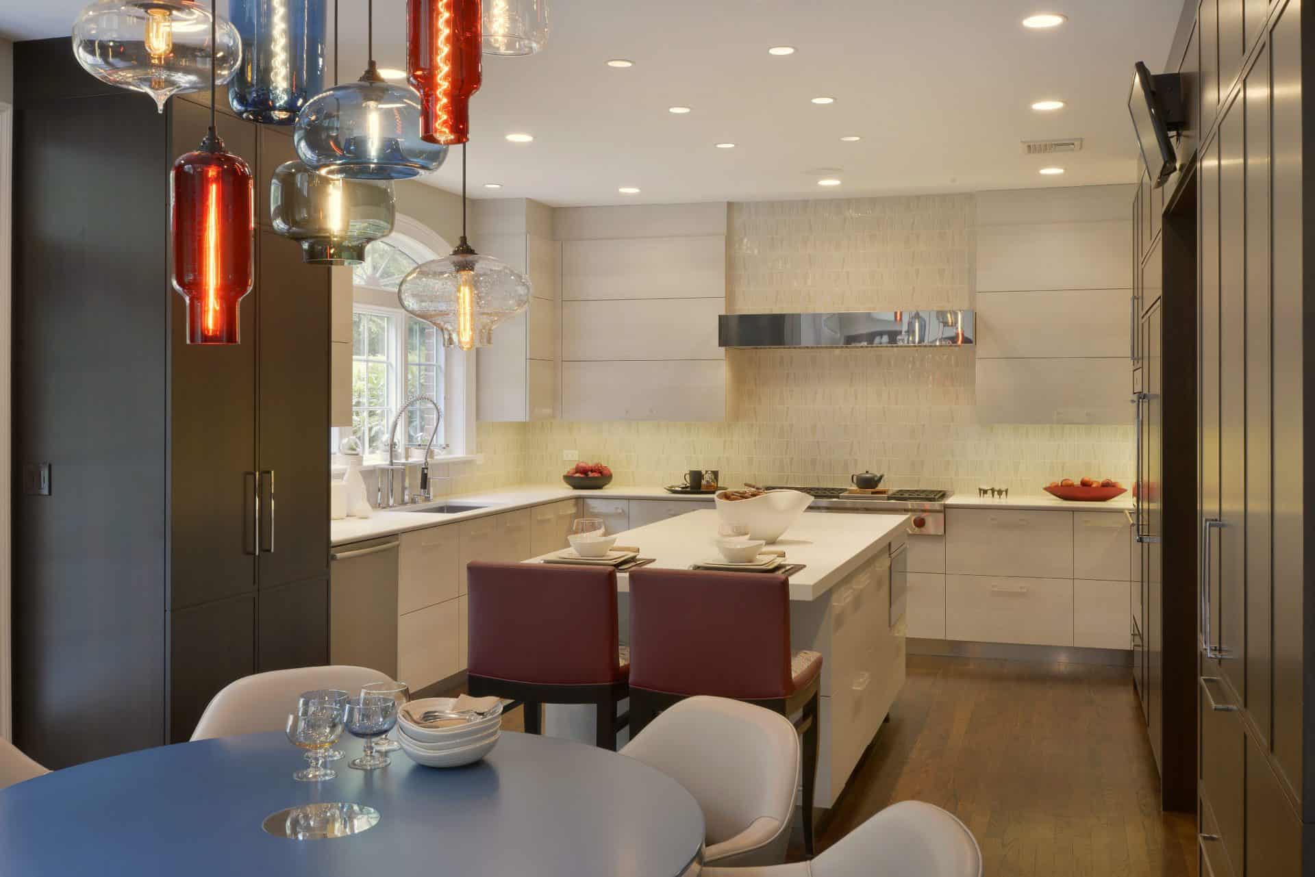 U-shaped Armonk kitchen features a center island and eat-in breakfast nook and fully custom, frameless Artcraft Cabinetry in a mix of flat panel with white textured laminate and shaker style in walnut. Red accents appear in the leather stools at the island and the pendant lights over the breakfast nook.