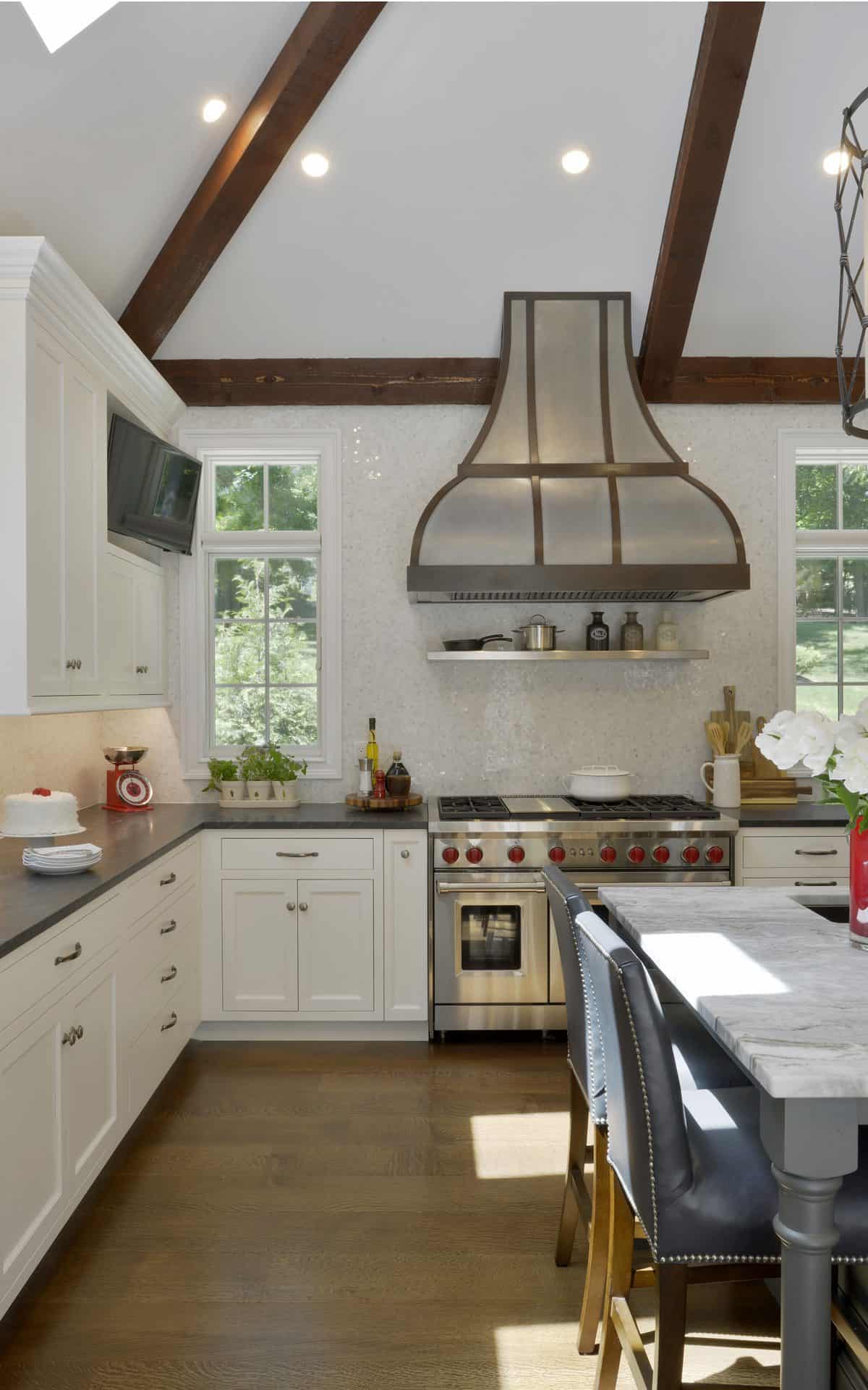 White classic kitchen features dark wood beams, elegant hoodvent, large windows and white Bilotta cabinetry.