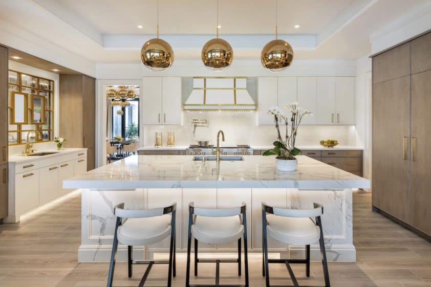 Dramatic white classic kitchen features custom Bilotta cabinets and range, expansive marble custom island, integrated appliances, and gold accents.