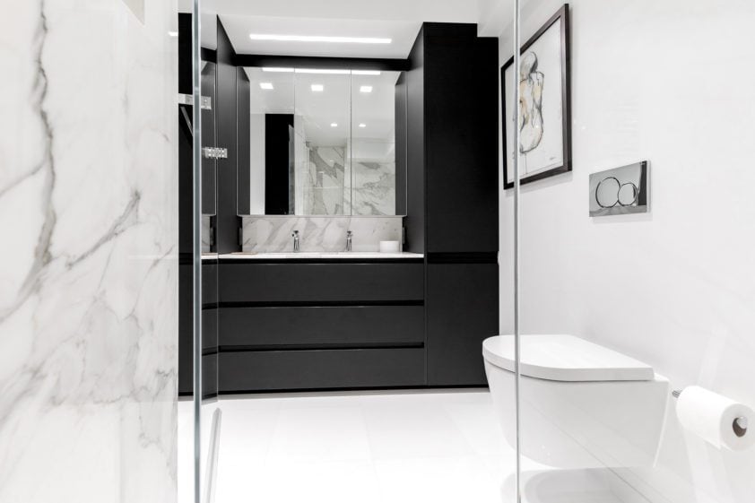 Dramatic bathroom features sleek black handle-less caninetry, marbled walls and modern bathroom appliances.