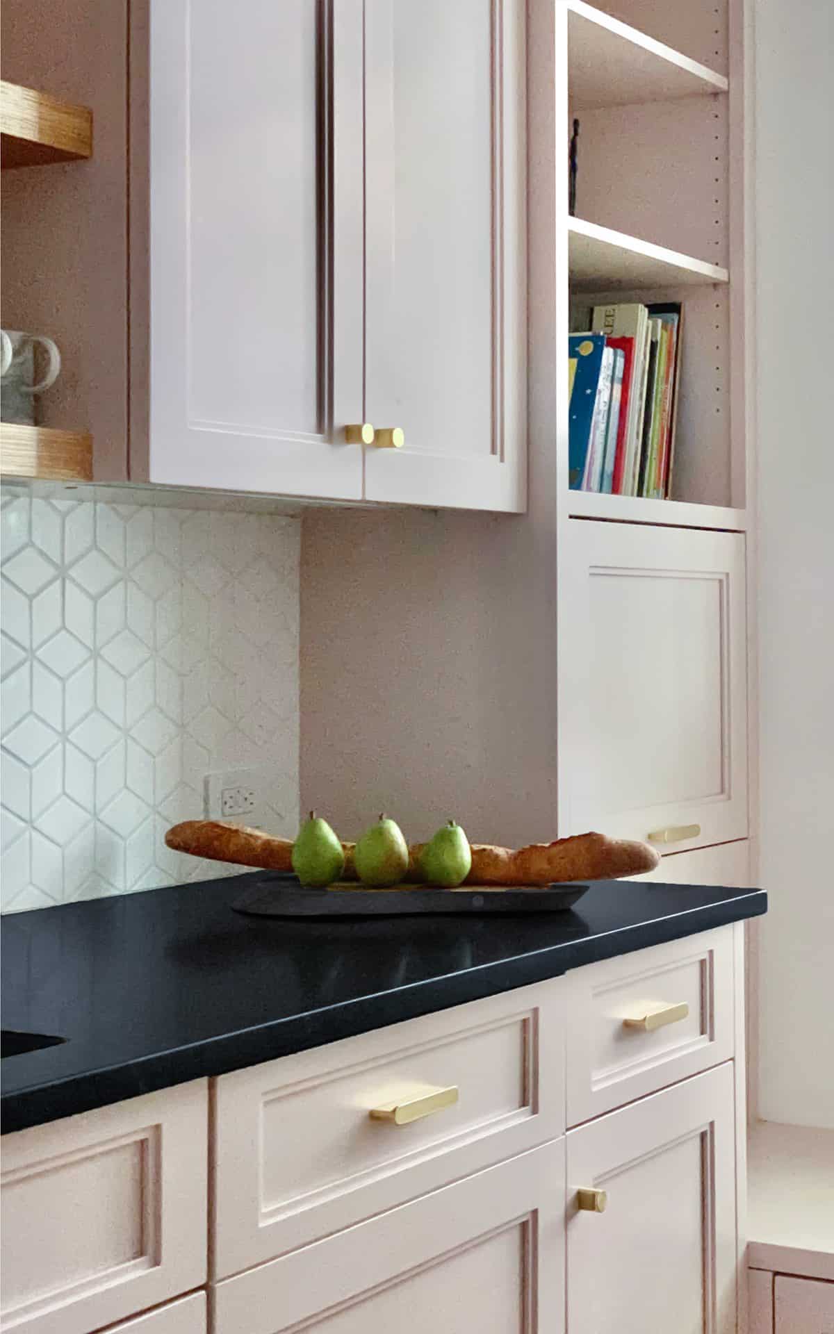 Kitchen with pale pink Bilotta custom cabinets, gold hardware, white backsplash and dark custom countertops.