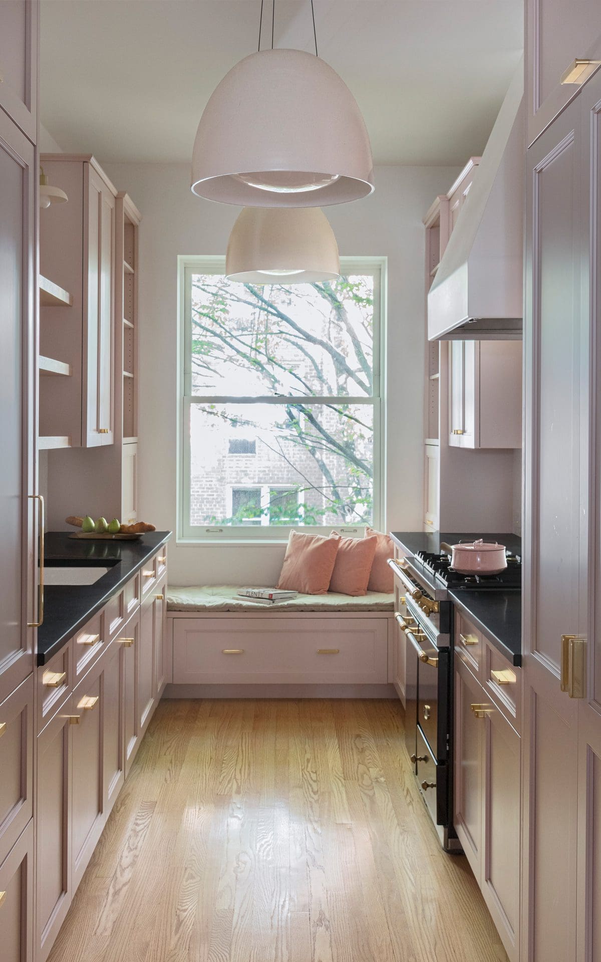 Classic kitchen features large window with window seat, pink cabinetry and dark countertops.