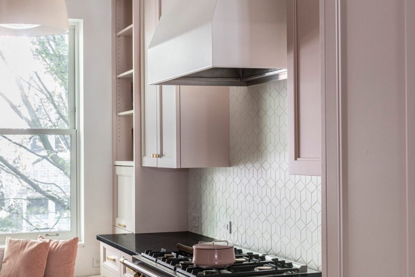 Classic kitchen with pale pink cabinetry, custom hood vent, classic white backsplash and large pendant lights.
