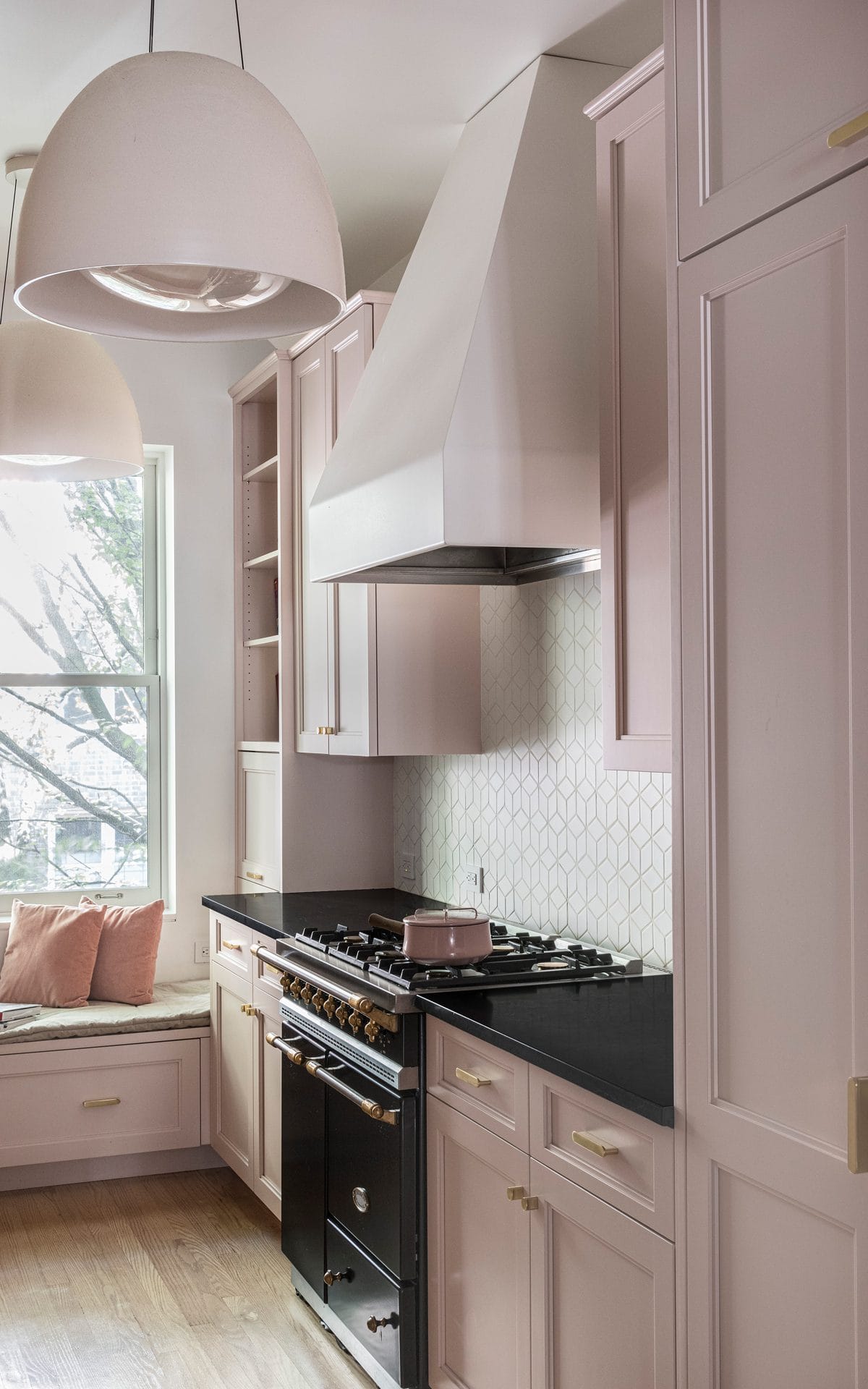 Classic kitchen with pale pink cabinetry, custom hood vent, classic white backsplash and large pendant lights.