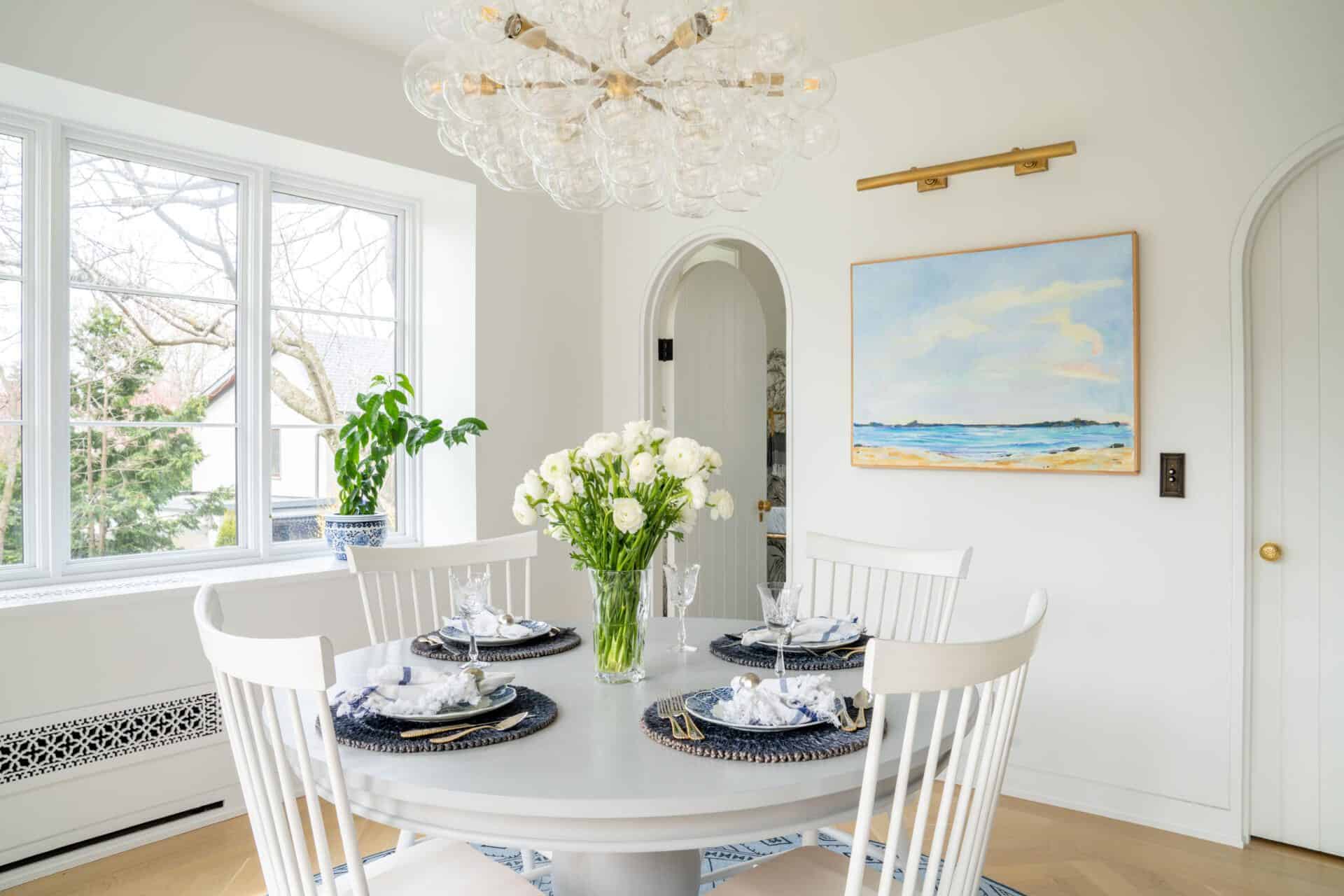 Dining area with white chairs, gray circular table, bubble-like light fixture and seascape painting on wall