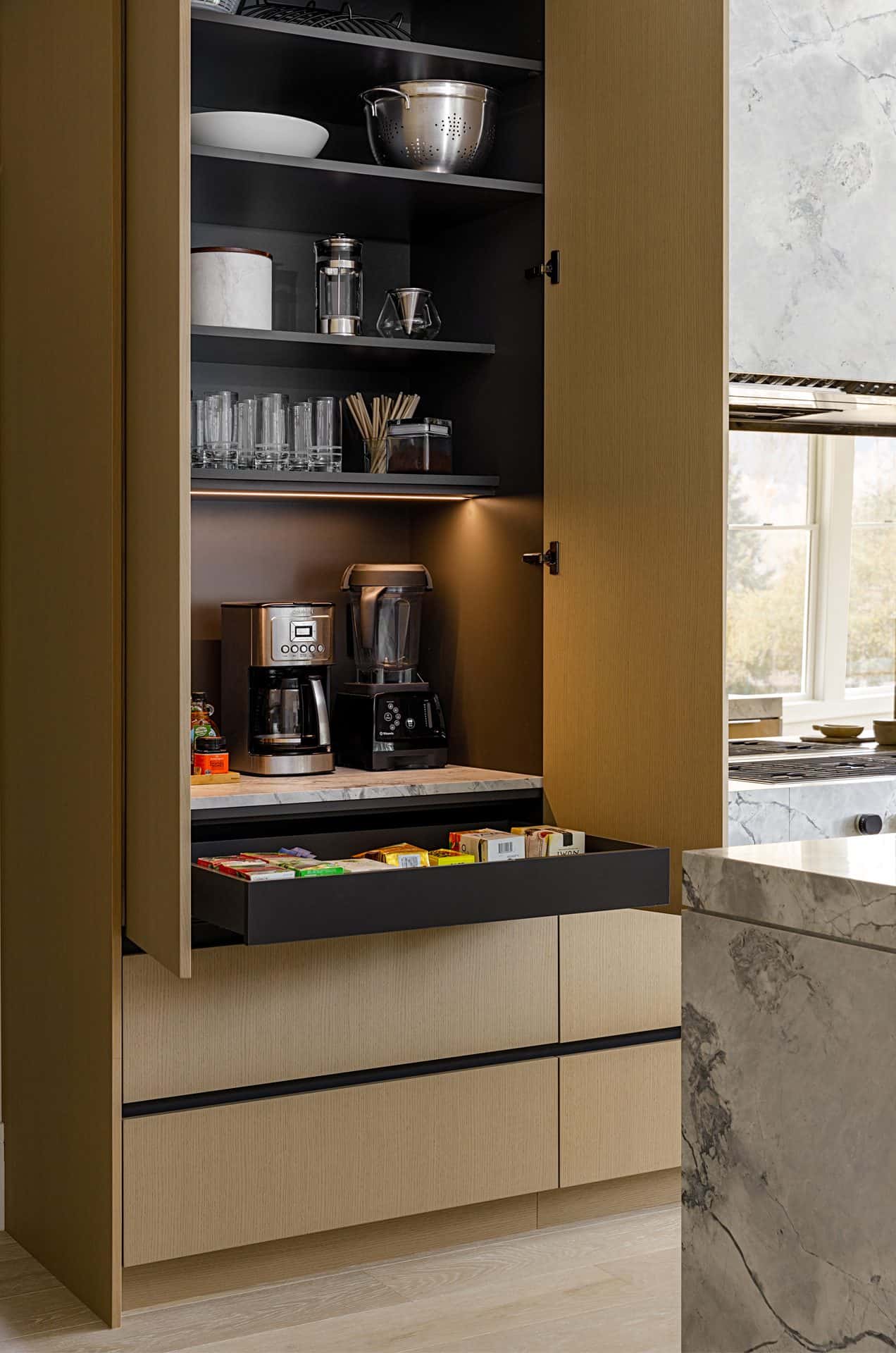 Close up of opened cabinet and drawer, next to range with polished stainless steel backsplash and with marble hood, countertops and waterfall edge island.