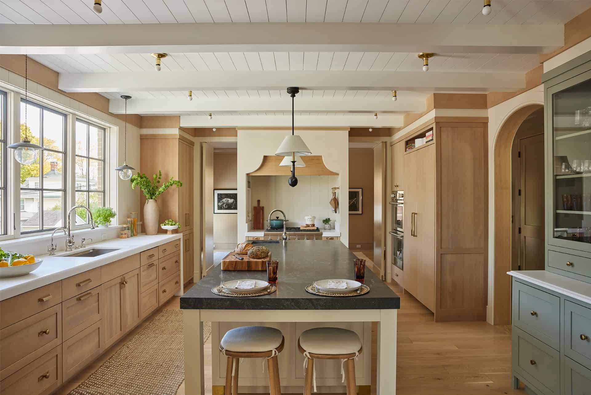 Overall view of kitchen with base oak cabinets on the left with a sink, long island with seating for two at the end, cooking nook in the back, paneled refrigerator and custom hutch on the right
